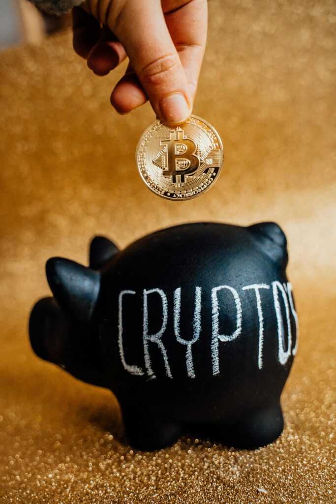 A hand places a Bitcoin into a piggy bank labeled 'Crypto', symbolizing savings and investment.